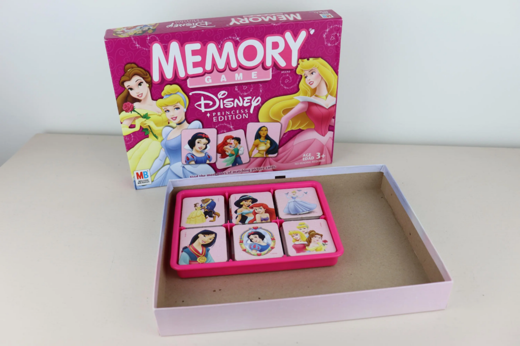 box that says "memory game: disney princess edition" and the box has an image of belle, Cinderella, and aurora, as well as pictures of 3 tiles one with snow white, one with ariel and Eric and one with Pocahontas.
in front of the box there is a 2x3 tray that holds the tiles from left to right:
row 1; pocahontas, aurora, jasmine and aladdin
row 2: beast, snow white and Prince Florian, and dopey.
under the tray the rule book peaks out on the table.