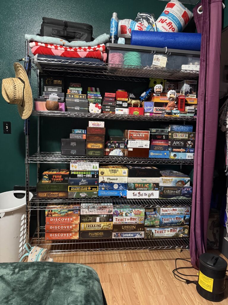 a shelf with significantly more games on it. on the top shelf there are blankets, crocheting supplies and various other object. on the side there is a hat hanging from the side.